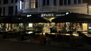 Der Außensitzbereich der Fasil Lounge Bar bei Nacht, mit Tischen und Stühlen unter großen schwarzen Schirmen, beleuchtet von warmen Lichtern und dekoriert mit Topfpflanzen vor einem modernen Gebäude.