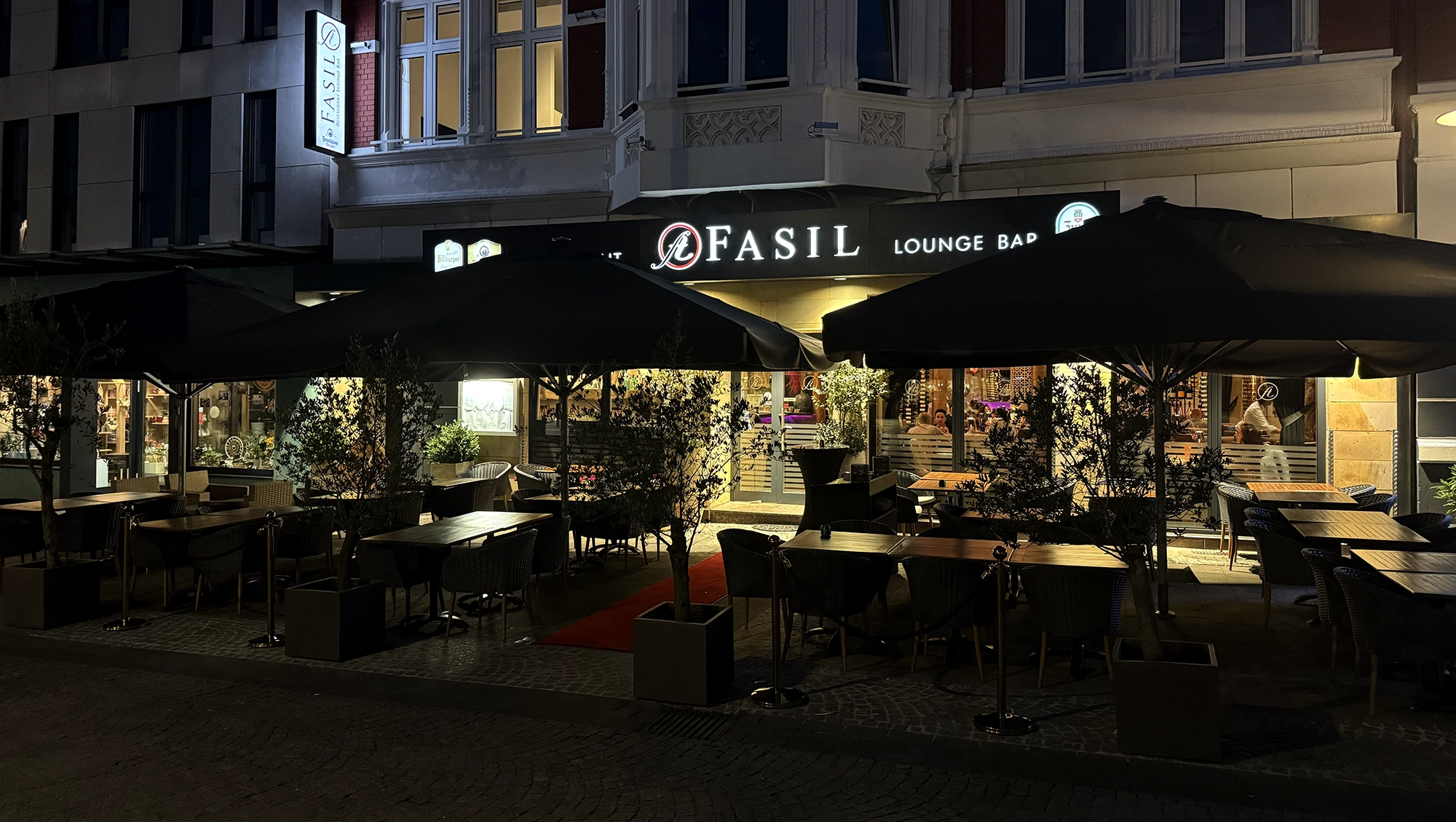Der Außensitzbereich der Fasil Lounge Bar bei Nacht, mit Tischen und Stühlen unter großen schwarzen Schirmen, beleuchtet von warmen Lichtern und dekoriert mit Topfpflanzen vor einem modernen Gebäude.