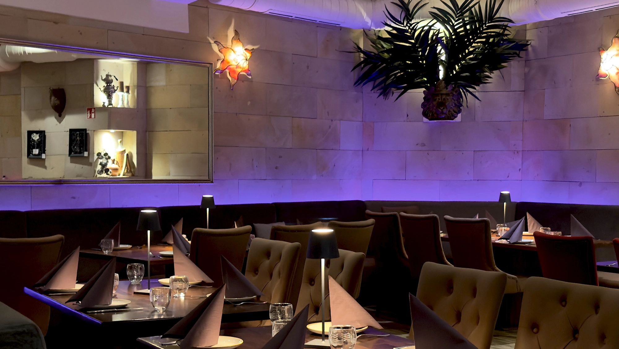 Ein modernes Restaurantinterieur mit ordentlich gedeckten Tischen, braunen Stühlen, gefalteten Servietten, Gläsern und kleinen Lampen. Sanftes violettes Licht und dekorative Wandbefestigungen schaffen ein gemütliches Ambiente. Ein großer Wandspiegel reflektiert einen Teil des Raums.