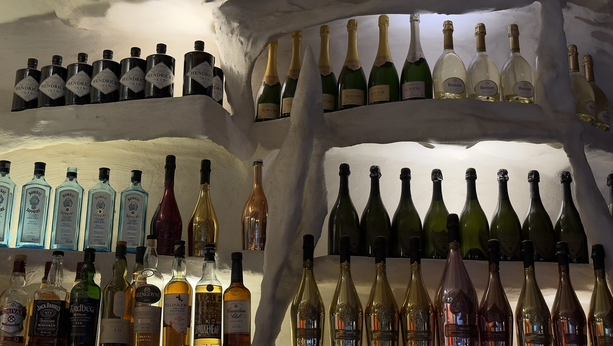 In einer weißen, höhlenartigen Wand sind Regale mit verschiedenen Spirituosen- und Champagnerflaschen in unterschiedlichen Formen und Farben eingelassen, die alle von einer sanften, warmen Beleuchtung angestrahlt werden.