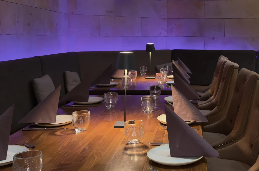 Ein moderner, für eine Gruppe gedeckter Restauranttisch mit gefalteten Servietten, Tellern und Gläsern. Sanftes violettes Licht und stilvolle Stühle mit hoher Lehne schaffen eine elegante, einladende Atmosphäre.