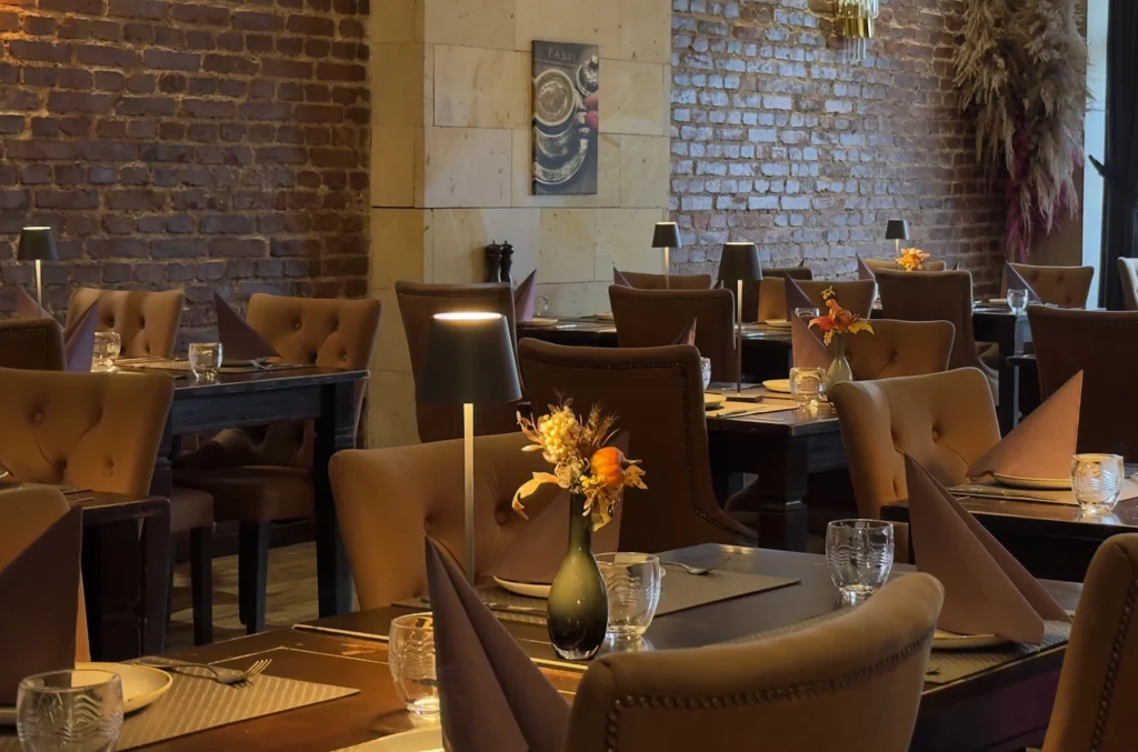 Ein gemütlicher Restaurantbereich mit braunen Lederstühlen, dunklen Holztischen mit Gläsern, Servietten und kleinen Blumenvasen. Warme Beleuchtung und freiliegende Backsteinwände schaffen eine einladende Atmosphäre.