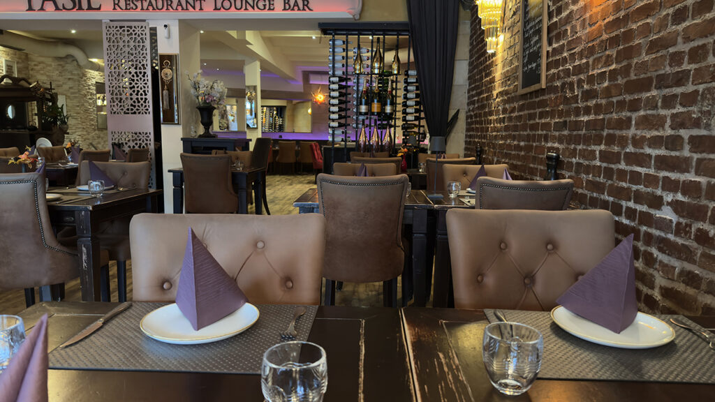 Elegantes Restaurantinterieur mit braunen getufteten Stühlen, Tischen mit Tellern, Gläsern und lila Servietten. Backsteinwände, warme Beleuchtung und ein Weinregal schaffen eine gemütliche, einladende Atmosphäre. Die Bar ist im Hintergrund zu sehen.