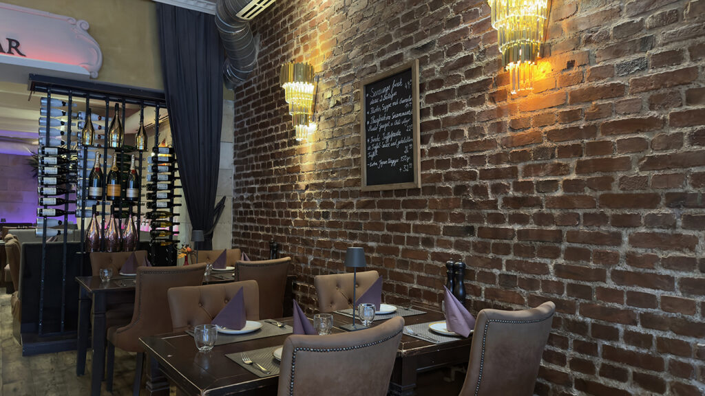 Ein gemütliches Restaurant mit freiliegenden Backsteinwänden, eleganten braunen Tuftingstühlen, ordentlich gedeckten Tischen, goldenen Hängelampen und einer Speisekarte aus Kreide. Im Hintergrund ist ein Weinregal zu sehen.