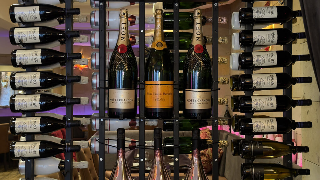 In einem Weinregal stehen mehrere Flaschen Rot-, Weiß- und Roséwein, in der Mitte zwei große Champagnerflaschen von Moët & Chandon und eine Flasche Veuve Clicquot. Im Hintergrund ist ein sanft beleuchtetes Restaurantinterieur zu sehen.