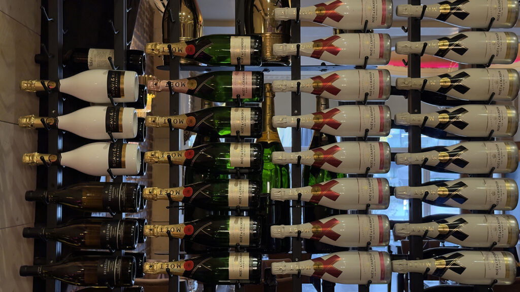 Champagner- und Sektflaschen, horizontal auf einem schwarzen Metall-Weinregal angeordnet, in Reihen gestapelt, mit Goldfolie um die Hälse und verschiedenen sichtbaren Etiketten. Der Hintergrund ist sanft beleuchtet.