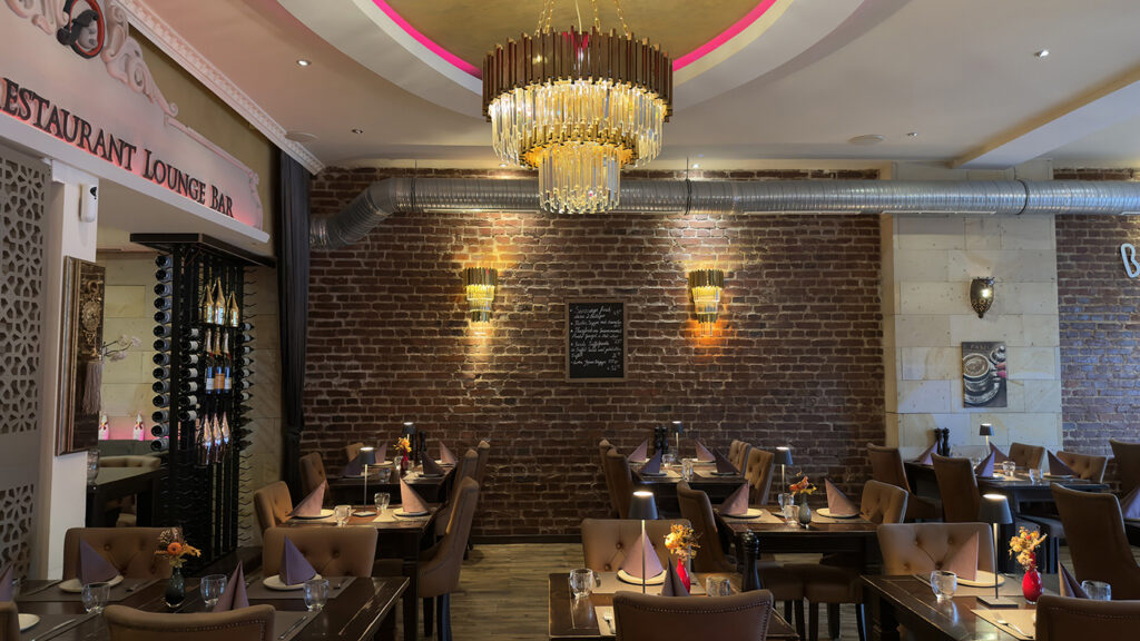 Ein gemütliches Restaurant mit freiliegenden Ziegelwänden, braunen, getufteten Stühlen, gedeckten Tischen mit Blumen, einem großen Kronleuchter, Wandleuchten und modernen Lüftungsschächten. An der Wand hängt eine Speisekarte aus Kreide.