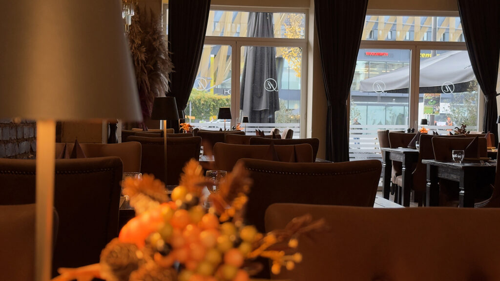 Ein gemütliches, warm beleuchtetes Restaurant mit braunen Stühlen und Tischen, dekoriert mit herbstlichen Tischdekorationen. Große Fenster im Hintergrund geben den Blick auf ein modernes Gebäude und Sonnenschirme im Freien frei.