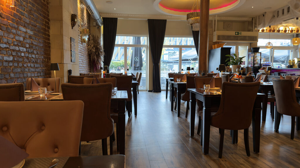 Ein gemütliches, modernes Restaurant mit braunen Ledersesseln, dunklen Holztischen für die Gäste, cremefarbenen Backsteinwänden, großen Fenstern mit schwarzen Vorhängen und warmer Raumbeleuchtung.