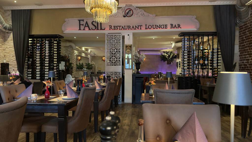 Elegantes Restaurantinterieur mit braunen Lederstühlen, Tischen mit gefalteten lila Servietten, Weinregalen, Zierpflanzen und einem Kronleuchter. Ein Schild mit der Aufschrift "FASIL Restaurant Lounge Bar" steht über einer Trennwand. Warme, stimmungsvolle Beleuchtung.