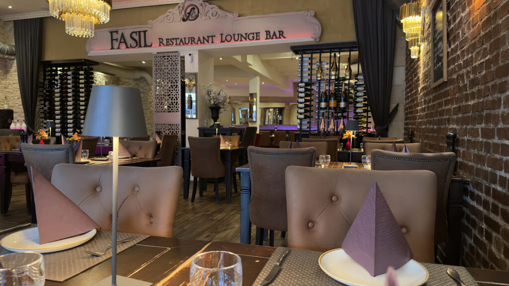 Elegantes Restaurantinterieur mit braunen Lederstühlen, ordentlich gedeckten Tischen mit gefalteten Servietten, Weingläsern und einem dekorativen Weinregal. Warme Beleuchtung und Kronleuchter schaffen eine gemütliche, gehobene Atmosphäre.