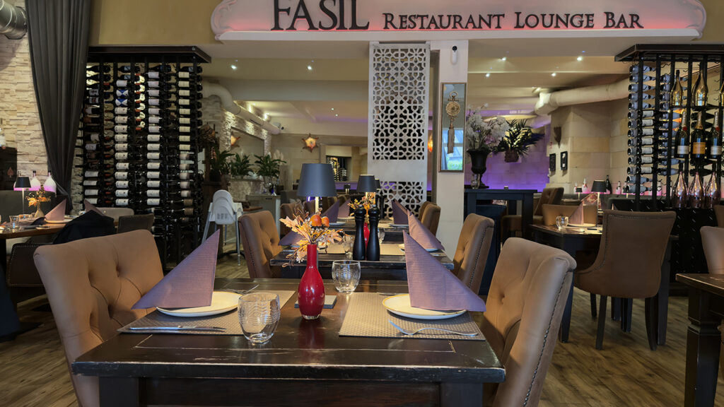 Elegantes Restaurantinterieur mit ordentlich gedeckten Tischen, gefalteten Servietten, Weingläsern, dekorativen Blumen und gedämpfter Beleuchtung. Weinregale und gemütliche, gepolsterte Stühle unterstreichen die einladende Atmosphäre. Auf einem Schild steht "Fasil Restaurant Lounge Bar".