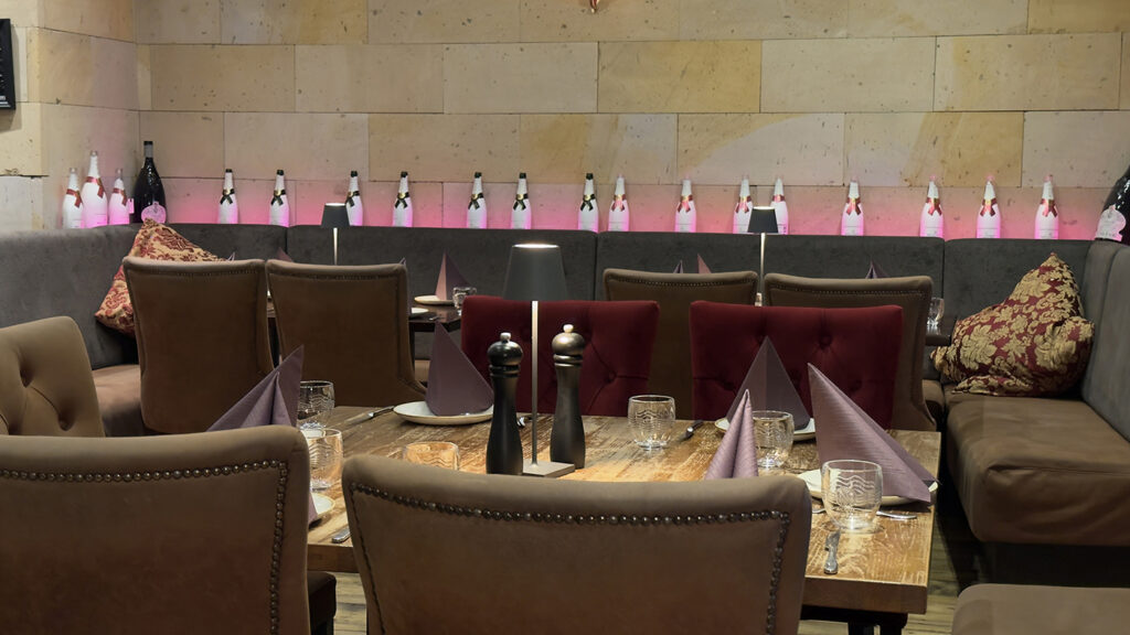 Ein gemütliches Restaurant mit braun gepolsterten Stühlen und Holztischen, die mit Gläsern, Tellern und gefalteten lila Servietten gedeckt sind. Dekorative Flaschen säumen ein Regal entlang der beigen Steinwand im Hintergrund.