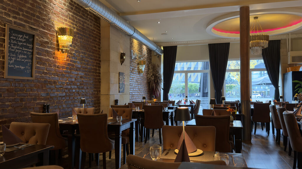 Ein gemütliches Restaurant mit braunen Lederstühlen, Holztischen zum Essen, freiliegenden Ziegelwänden, Kronleuchtern, großen Fenstern mit dunklen Vorhängen und einer Speisekarte an der Wand.