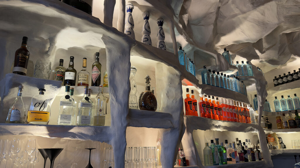 In den Regalen, die in eine weiße, höhlenartige Wand eingelassen sind, stehen Flaschen mit verschiedenen Spirituosen, darunter Wodka, Gin, Tequila und Liköre in verschiedenen Formen und Farben, und darunter sind die Gläser ordentlich angeordnet.