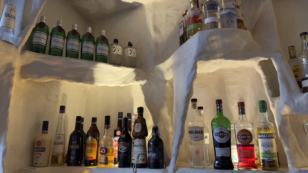 Flaschen mit verschiedenen alkoholischen Getränken stehen auf weißen, unregelmäßig geformten Regalen, die in eine strukturierte Wand eingelassen sind und von warmem Licht beleuchtet werden.