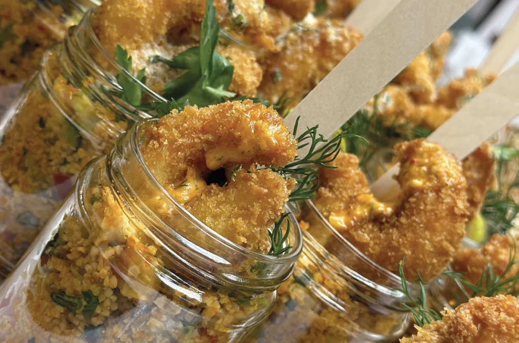 Nahaufnahme von panierten, gebratenen Garnelen, die in kleinen Gläsern serviert werden, garniert mit frischen Kräutern und Holzstäbchen, auf einem Bett aus Couscous oder Getreidesalat liegend.