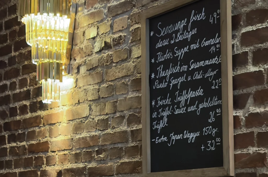 Eine Speisekarte mit handgeschriebenem deutschen Text hängt an einer Backsteinwand neben einem modernen Kronleuchter mit hängenden Glasstücken. Auf der Karte sind Gerichte und Preise in weißer Kreide aufgeführt.